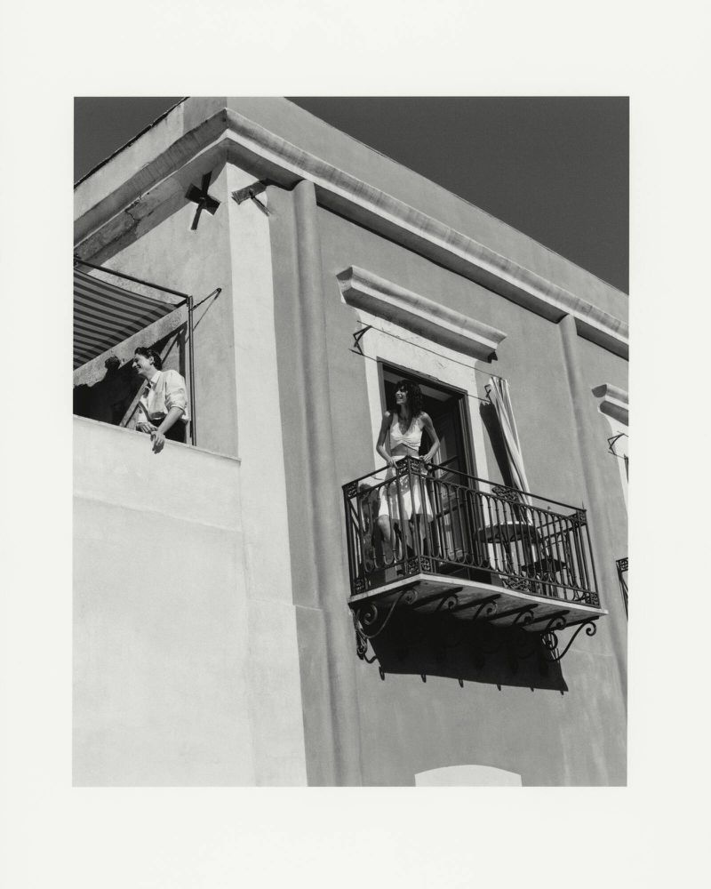 Two adults are standing on separate balconies of a multi-story building in a black-and-white architectural shot with a dark sky.