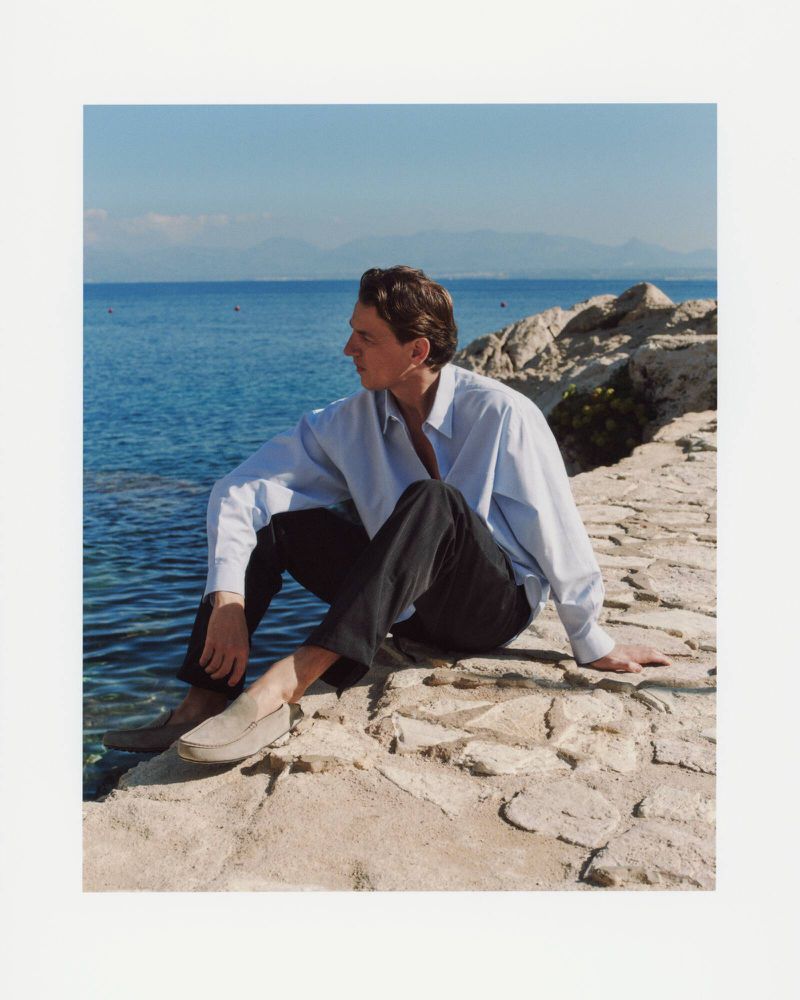 Adult male sits on a stone breakwater wall near the ocean with mountains in the distance on a sunny day. He wears a button-down shirt.