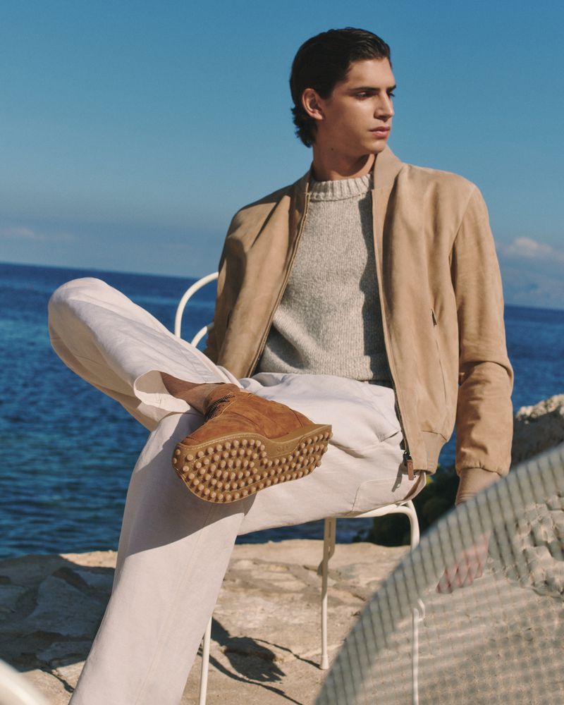Adult male in a beige jacket and pants is sitting on a chair with brown boots in front of a blue ocean on a sunny day.
