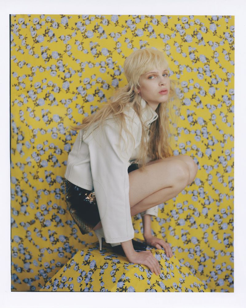 A fair-skinned model with blonde hair poses in a crouched position in front of a yellow backdrop patterned with small blue flowers.