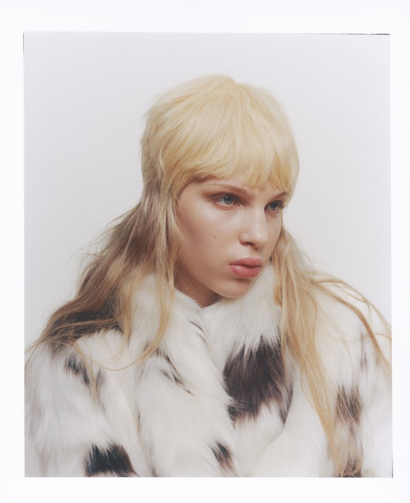 Close-up fashion shot of a blonde woman in a white and brown spotted fur coat against a bright white background in a photo studio.