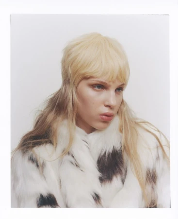 Close-up fashion shot of a blonde woman in a white and brown spotted fur coat against a bright white background in a photo studio.