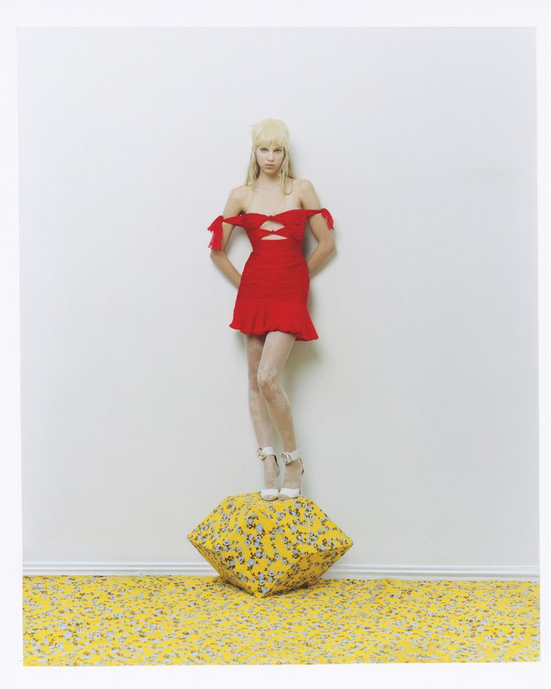 Woman in red dress stands on yellow floral cube in studio, posing against white wall. Full shot captures fashion and studio lighting.
