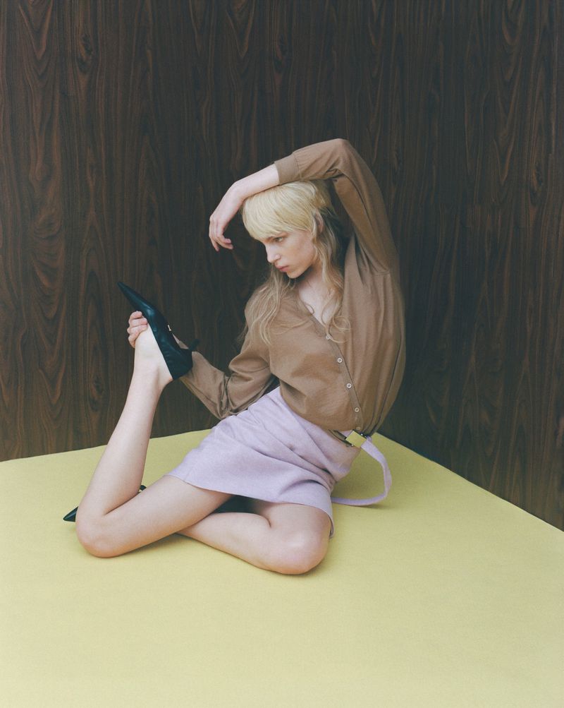Blonde model in a brown button-up shirt and lilac skirt posing dynamically on a yellow floor against a dark wood wall.
