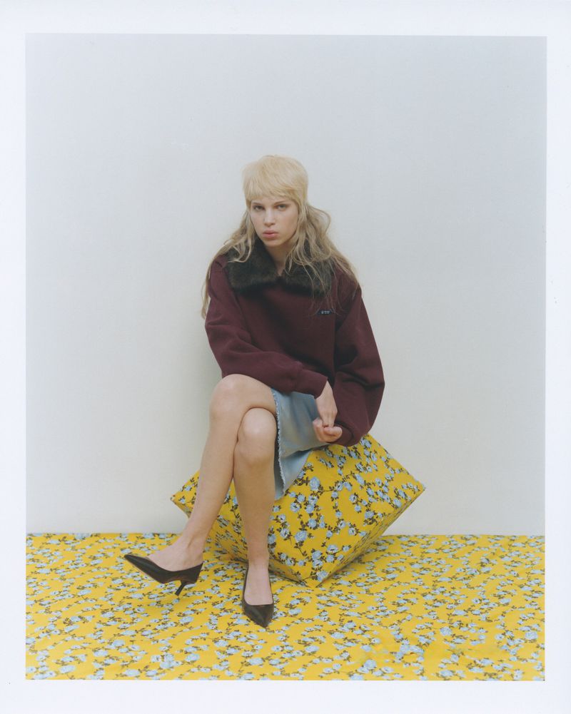 A blonde woman wearing a maroon sweater and denim skirt is sitting on a large yellow floral cushion in an indoor photo studio.