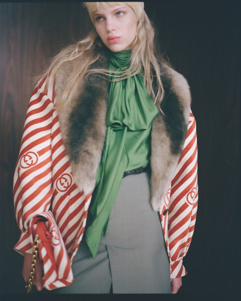 A blonde fashion model poses in a green blouse and patterned Gucci coat, set against a dark backdrop, emphasizing the textures and colors.