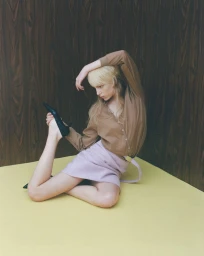 Blonde model in a brown button-up shirt and lilac skirt posing dynamically on a yellow floor against a dark wood wall.