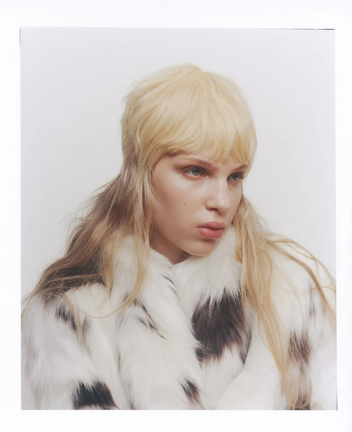Model with blonde shaggy haircut wearing a white and black faux fur coat against a white background.