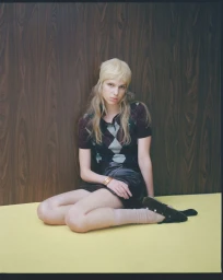 A blonde woman sits on a yellow floor against a dark wood wall wearing an argyle polo sweater, dark mini skirt, and clogs.