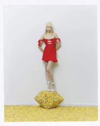 Blonde model wearing a red off-shoulder cutout mini dress standing on a yellow floral geometric block against a white wall.
