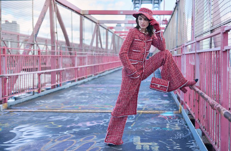 A woman in a red tweed suit and matching hat poses on a bridge, showcasing fashion and urban style with the city in the background.