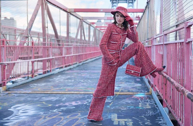 A woman in a red tweed suit and matching hat poses on a bridge, showcasing fashion and urban style with the city in the background.