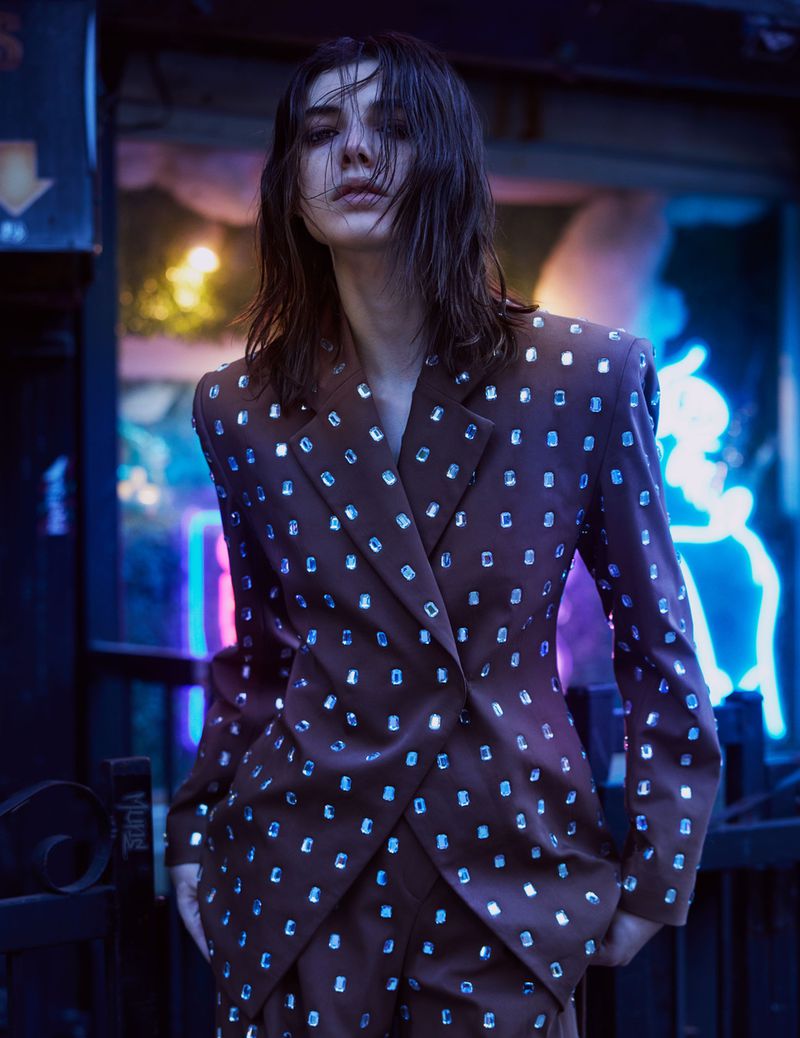 Woman in brown suit jacket with shimmering squares posing against blue neon-lit background. Fashion editorial style, wet hair, studio lighting.