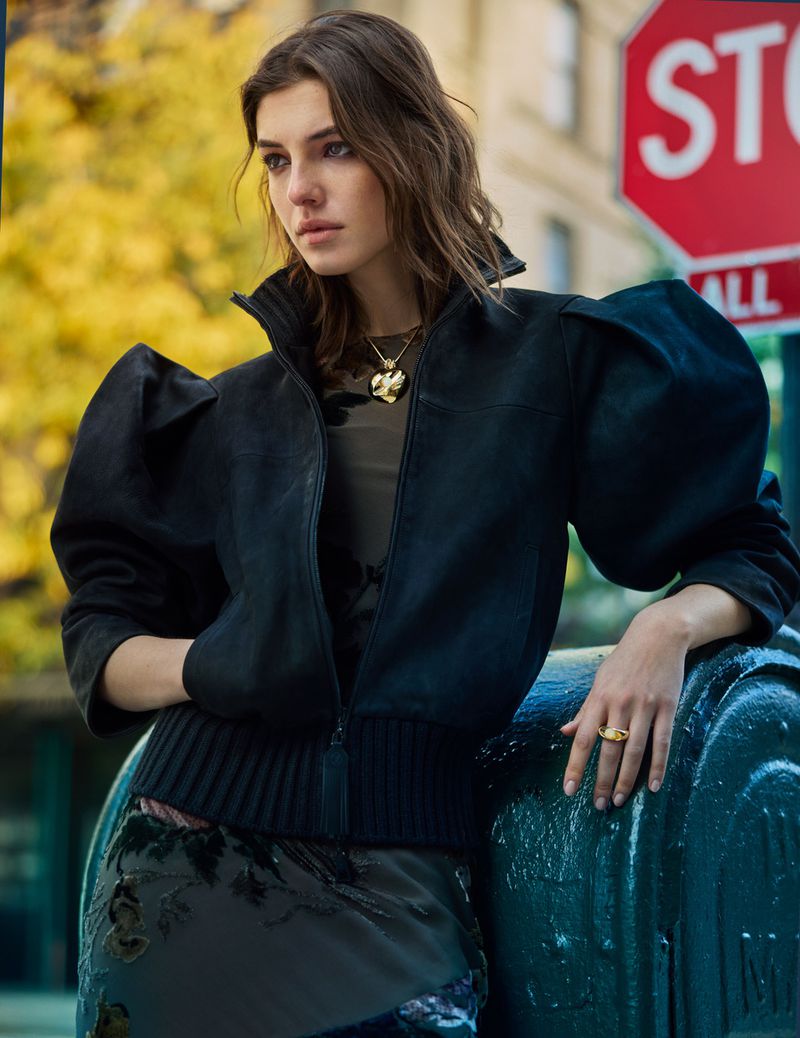 Model Carla Rey wears a black puff-sleeve bomber jacket and gold heart necklace while leaning against a dark street fixture outdoors.