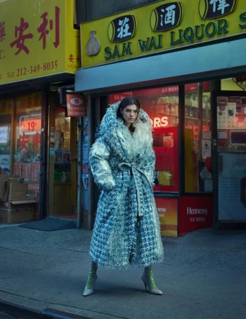Woman in oversized blue coat stands in front of a liquor store with Chinese signs, wearing green high-heeled boots in an urban setting.