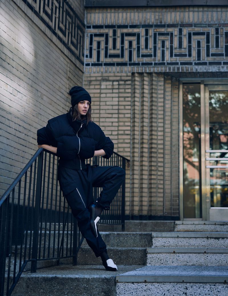 Woman in a black puffer jacket, beanie, and trousers leaning against metal railing on steps next to a brick building with decorative brickwork.