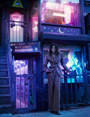 A woman poses in a brown and white polka dot suit, standing in front of a building with neon signs and metal gates in an urban environment.