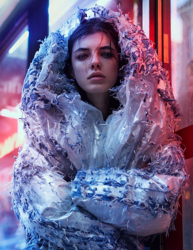 Portrait of a young woman with blue eyes wearing an elaborate hooded translucent plastic garment with blue accents, against a blurred background.