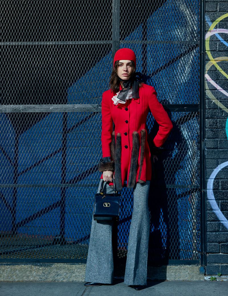 A woman in a red coat, hat, and gray pants poses with a black handbag in front of a blue metal grate with graffiti on a sunny day.