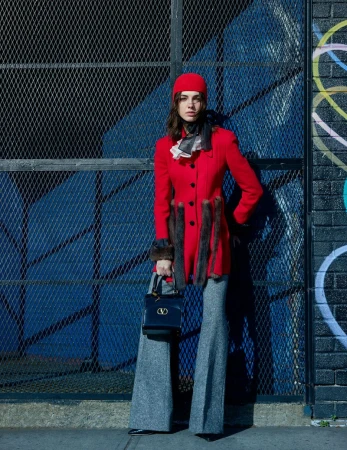 A woman in a red coat, hat, and gray pants poses with a black handbag in front of a blue metal grate with graffiti on a sunny day.