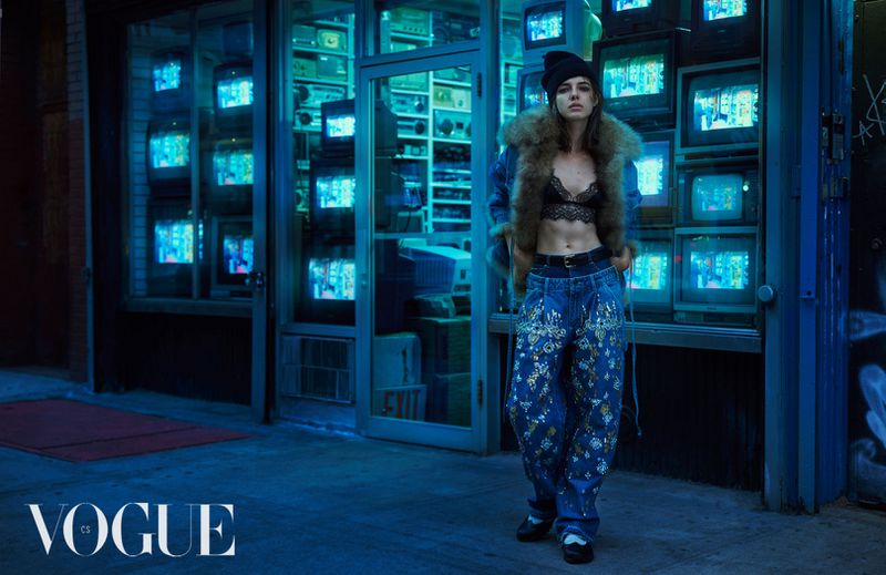 A model in a lace top and embroidered jeans stands on a city street in front of a vintage TV shop in a Vogue editorial shot.