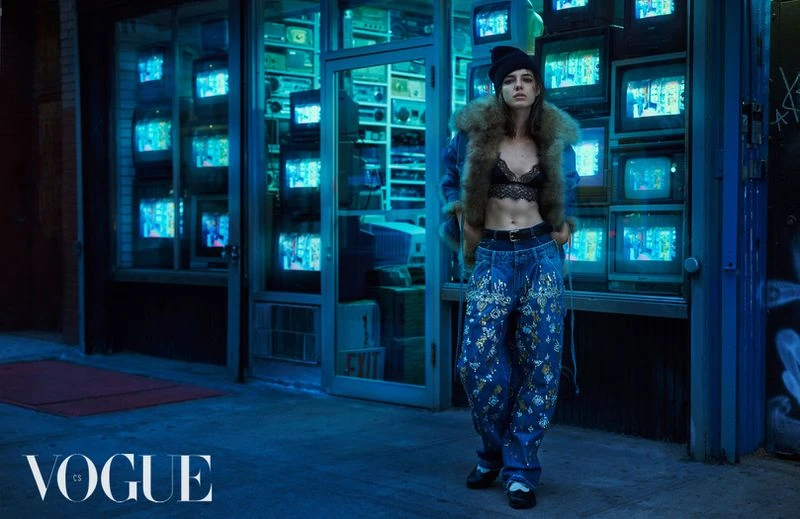 A model in a lace top and embroidered jeans stands on a city street in front of a vintage TV shop in a Vogue editorial shot.