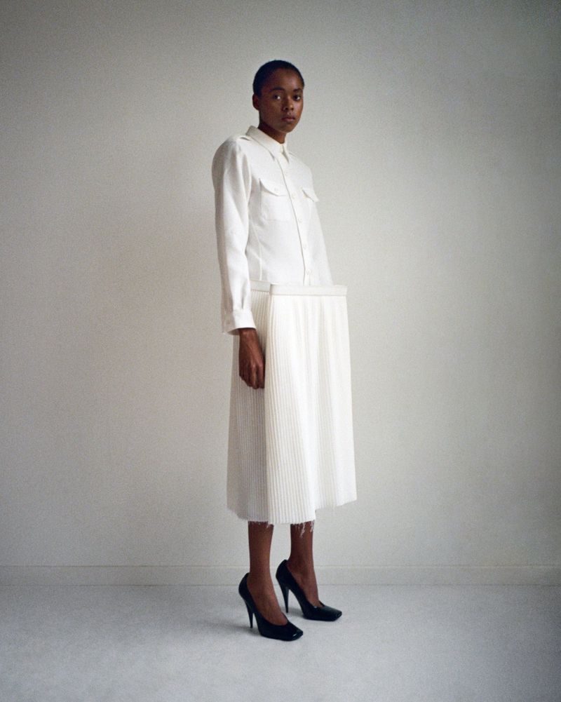 Young woman in a white button-down shirt and pleated skirt poses in black high heels against a stark white wall in a studio setting.