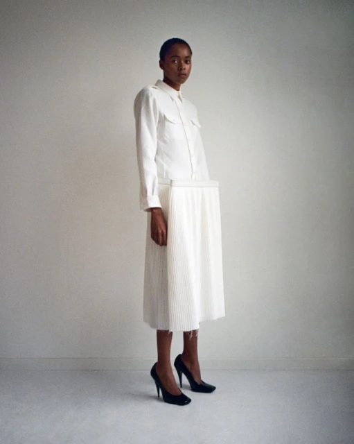 Young woman in a white button-down shirt and pleated skirt poses in black high heels against a stark white wall in a studio setting.