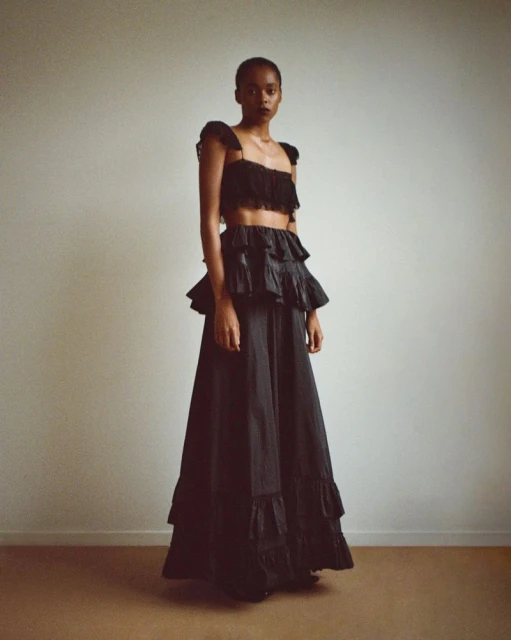 Fashion model in black two-piece outfit poses against a neutral wall, showcasing a stylish studio portrait with soft, diffused lighting.