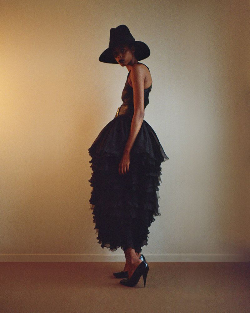 Model Nyla Singleton wearing a voluminous tiered black dress, wide-brim black hat, and black stiletto pumps against a warm wall.