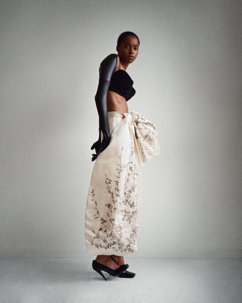 A striking studio shot features a Black female fashion model in a black top, dramatic gloves, and an ornate patterned skirt with black heels.