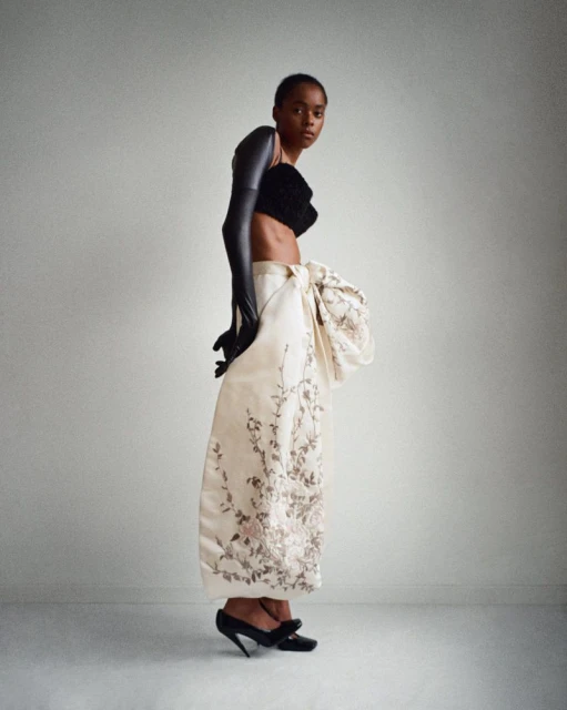 A striking studio shot features a Black female fashion model in a black top, dramatic gloves, and an ornate patterned skirt with black heels.