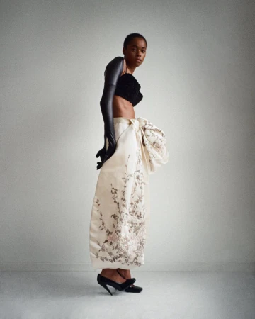 A striking studio shot features a Black female fashion model in a black top, dramatic gloves, and an ornate patterned skirt with black heels.