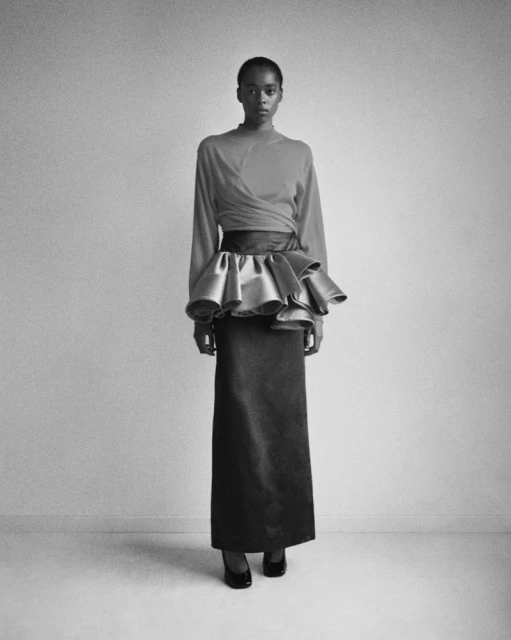 A black and white full shot presents a slender black female model in a high-waisted skirt and draped top, set against a plain wall.