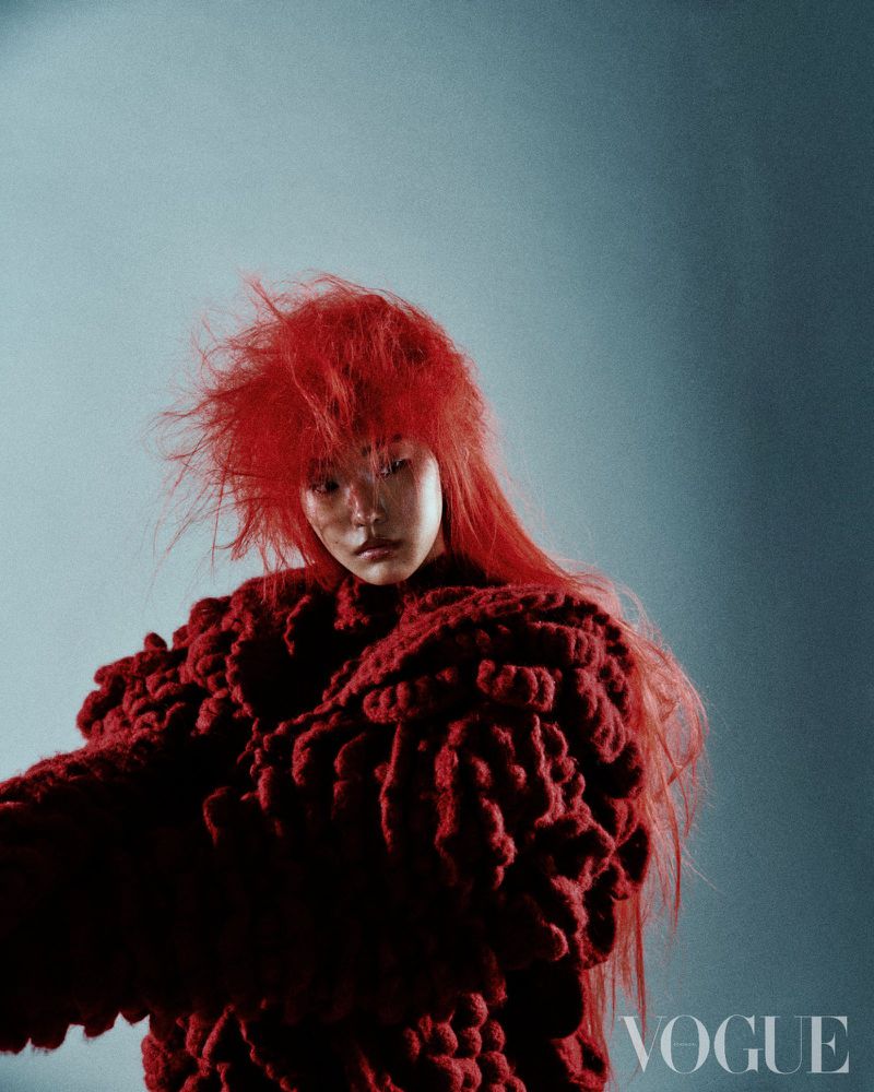 Fashion editorial portrait of a woman with red hair wearing a textured red sweater against a mottled blue studio background.