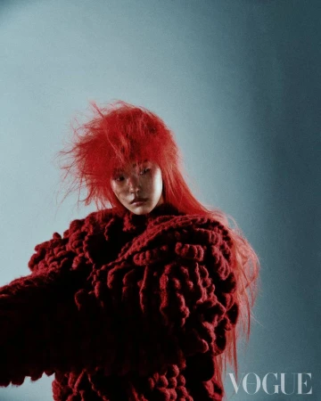 Fashion editorial portrait of a woman with red hair wearing a textured red sweater against a mottled blue studio background.