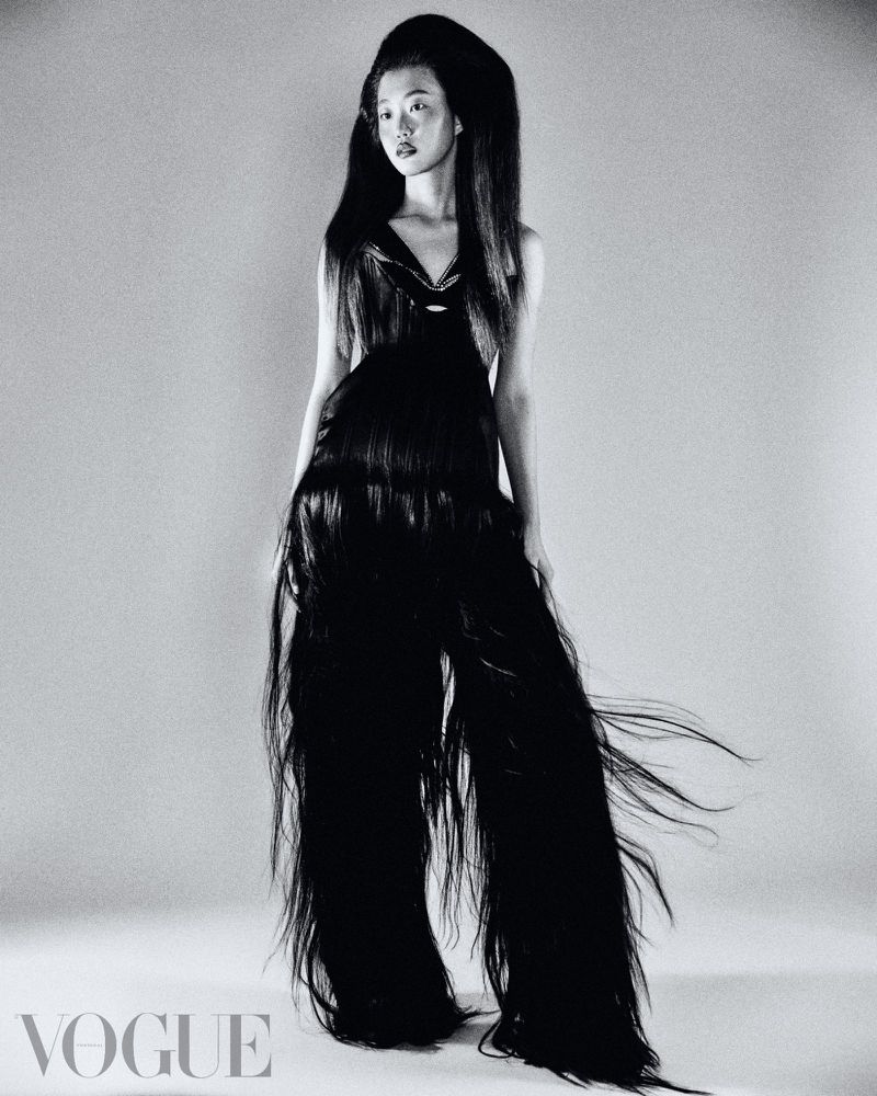 A model with long black hair poses in a studio, wearing a black dress made of hair, for a fashion editorial shot on a gray background.