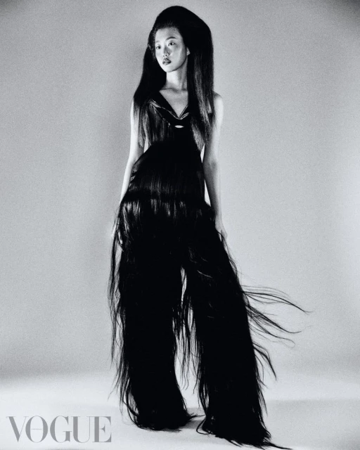 A model with long black hair poses in a studio, wearing a black dress made of hair, for a fashion editorial shot on a gray background.