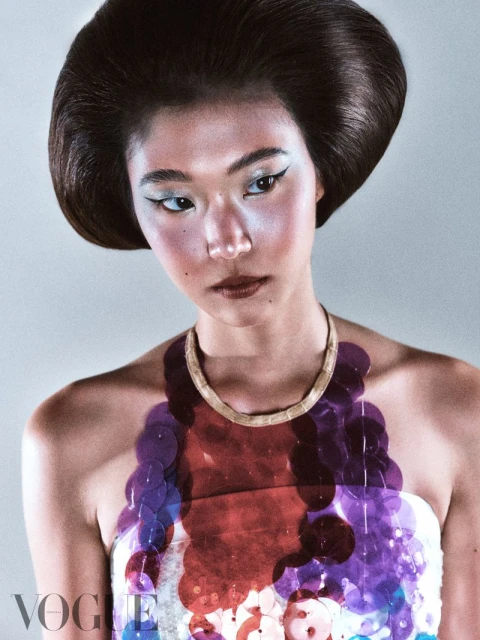 Fashion shot of an Asian woman with elaborate updo hairstyle and makeup, wearing a colorful sequin top, in front of a grey backdrop.