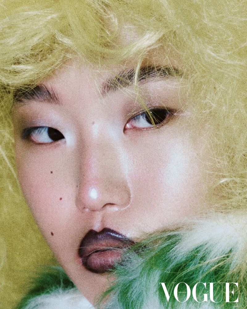 Close-up portrait of an Asian female model with dark lipstick and yellow textured hair. Vogue logo is visible at the bottom right.