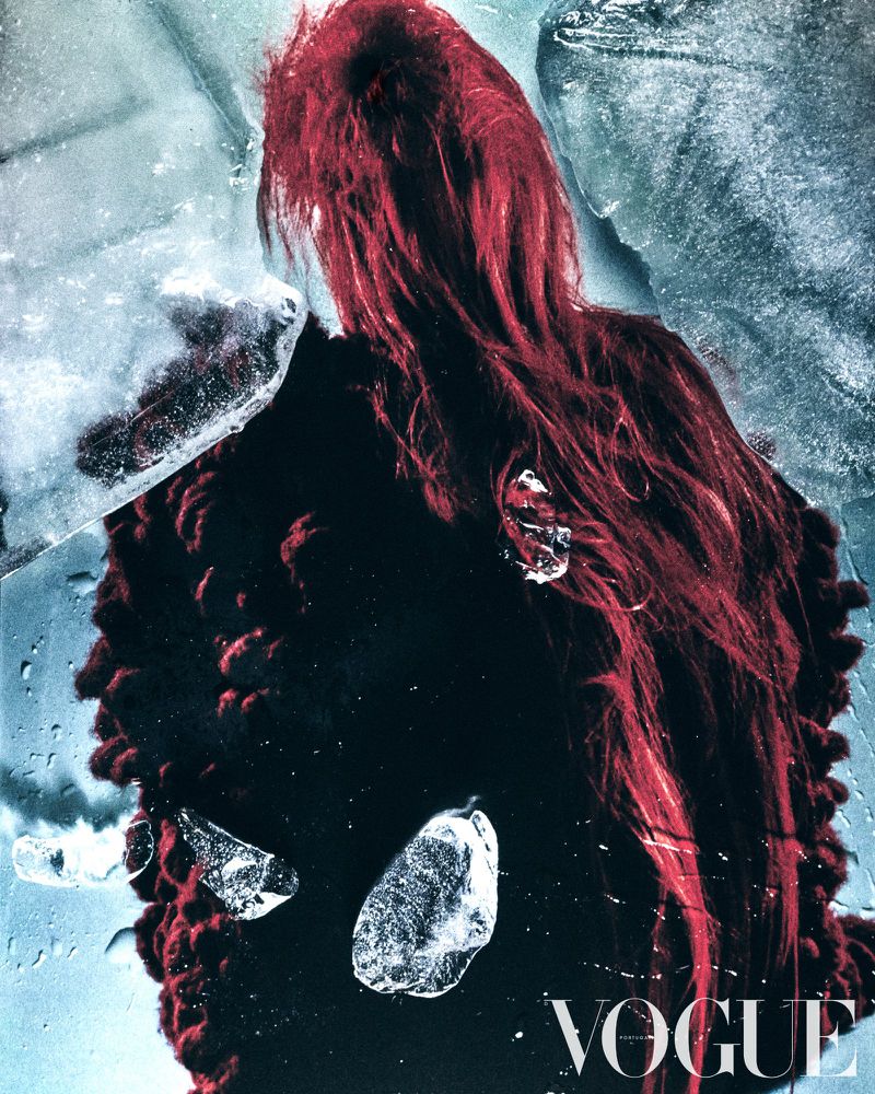 Fashion editorial shot features a person with red hair in a fuzzy black coat, ice shards, and Vogue magazine logo. Moody, cool-toned image.