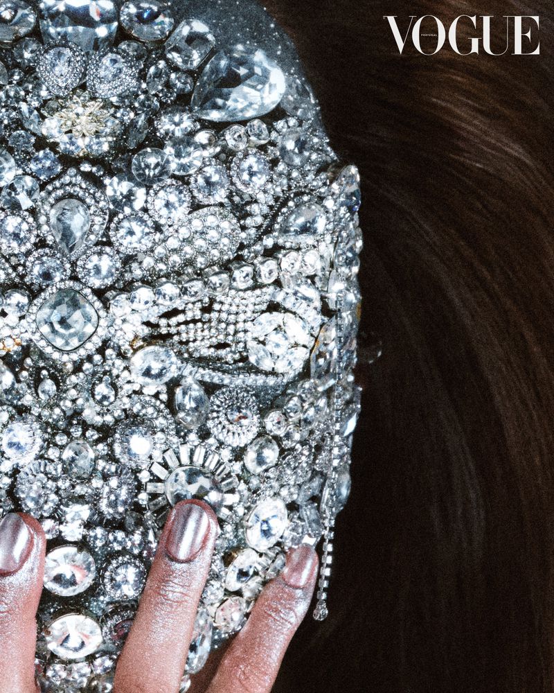 Close-up of a model's face covered in silver jewels and gems, posing for a Vogue magazine cover; dark brunette hair visible on right.