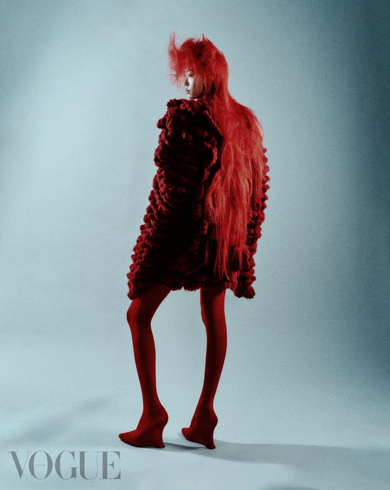 Fashion shoot of an adult female model in a red knit outfit with matching tights and shoes, posing in a studio against a light blue background.