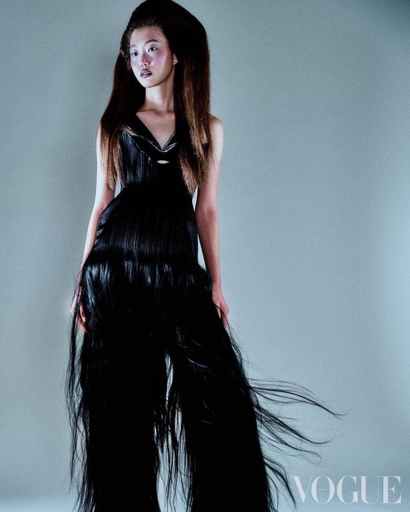 Model with voluminous dark hair wearing a black fringe jumpsuit against a grey seamless background