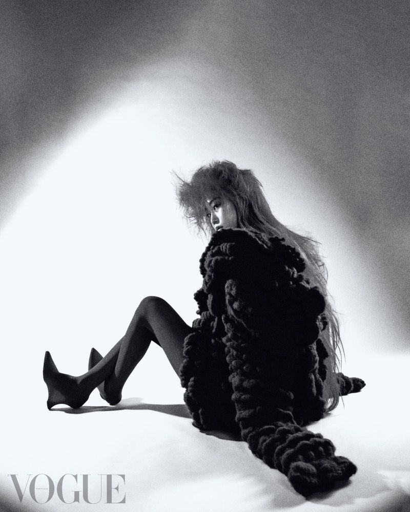 Fashion editorial photo of a woman in an avant-garde outfit sitting in a photo studio wearing stockings with a 'Vogue' logo visible.