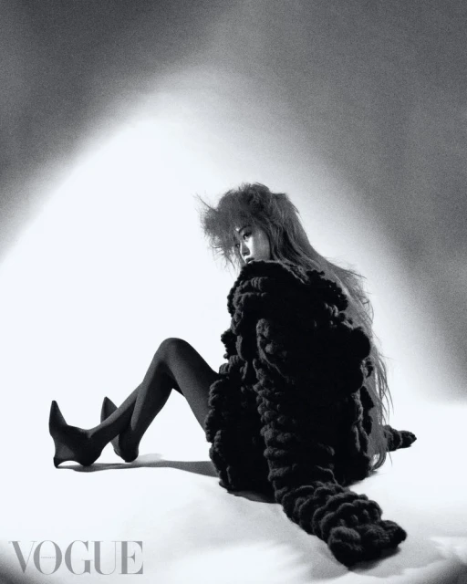 Fashion editorial photo of a woman in an avant-garde outfit sitting in a photo studio wearing stockings with a 'Vogue' logo visible.