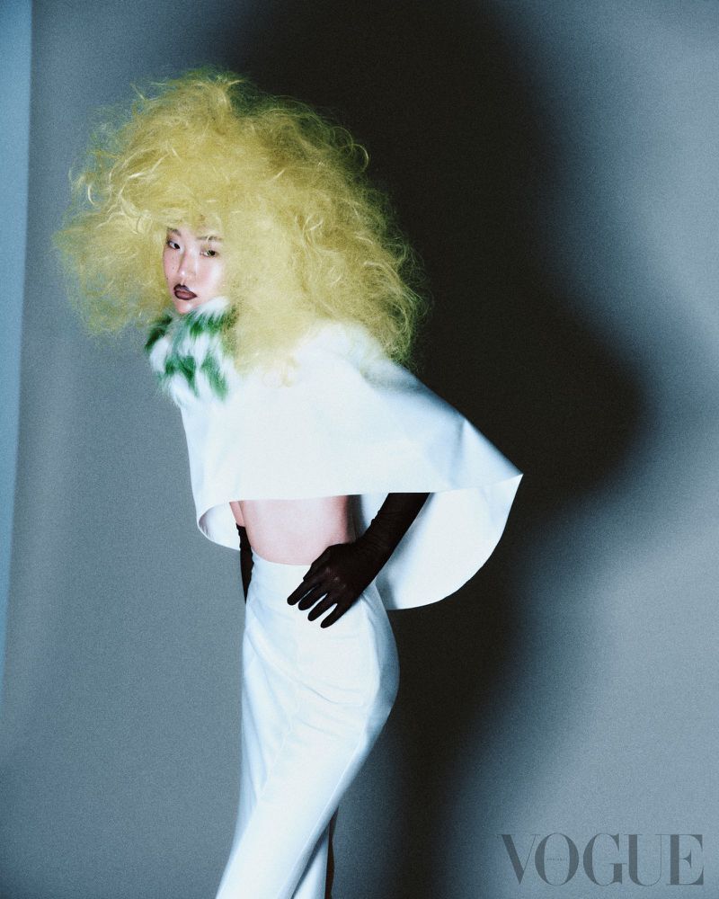 Fashion editorial image of a woman in white pants and top with a green and white fur collar, wearing a large blonde afro hairstyle.