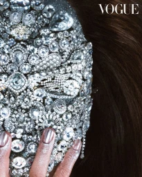 Close-up portrait of a face fully encrusted with sparkling crystal jewelry and diamonds with a silver-manicured hand touching the chin.