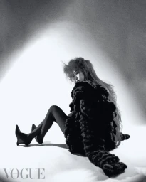 Monochrome fashion editorial image of a woman sitting on a white seamless floor wearing a textured dark coat and tights.