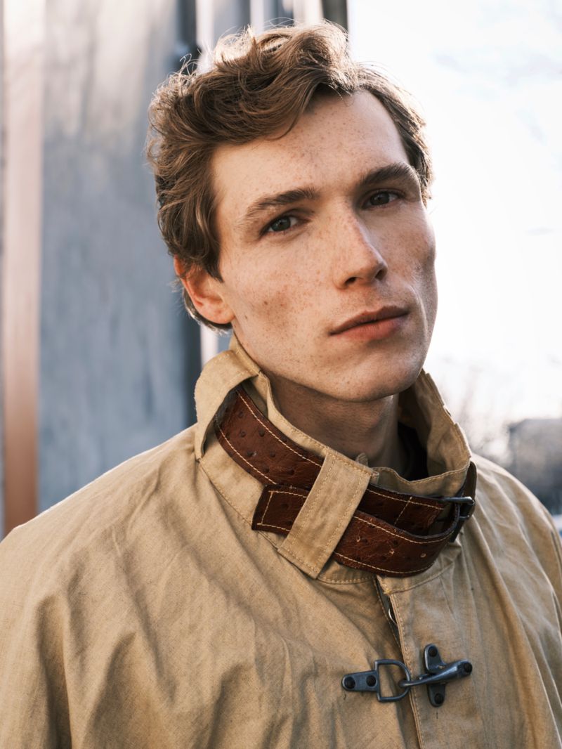Portrait of a young man with wavy red hair wearing a beige jacket with a brown leather collar, captured in a close-up shot.